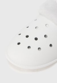 Vitt slip-on skor med cirkulära ventilationshål och mjukt fluffigt foder synligt vid hälområdet mot en vit bakgrund.