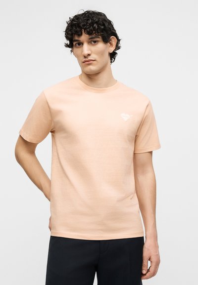 Νέος άνδρας με σγουρά σκούρα μαλλιά, που φοράει ροδακινί κοντομάνικη μπλούζα και μαύρο παντελόνι, στέκεται με το ένα χέρι πίσω από την πλάτη.