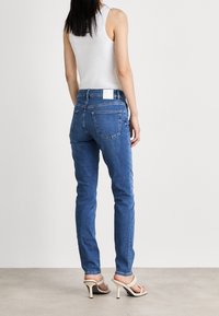 Blå denimjeans med slim fit, femficksdesign och en vit logotyp på bakre midjan. Kombination med en ljus linne och klackar.