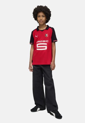 Maillot de football rouge et noir à manches courtes, grand numéro 5 blanc, et logo. Assorti à un jean noir à jambes larges et des chaussures de sport noires.