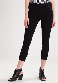 Černé zkrácené džíny s přiléhavým střihem a bočními knoflíkovými detaily, v kombinaci s černými sandály na podpatku s otevřenou špičkou. Pás je pokryt proužkovaným topem.