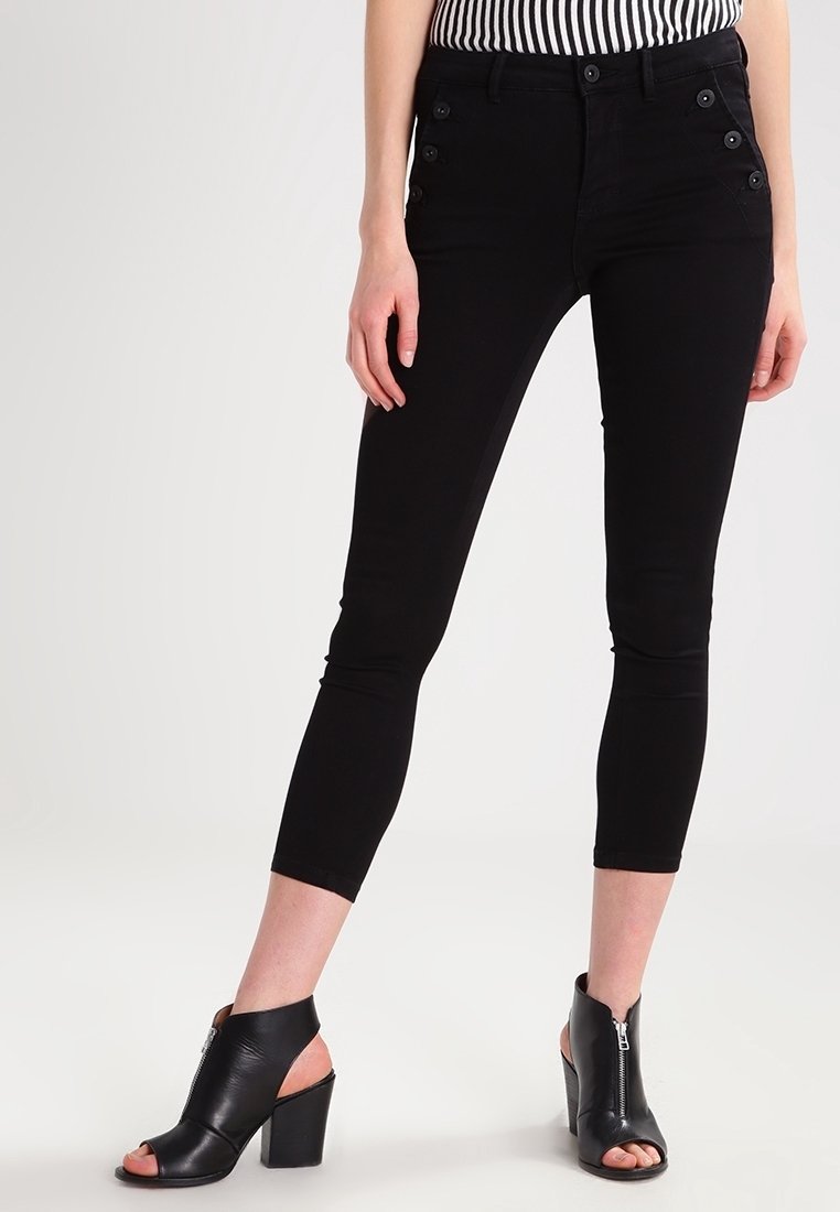 Černé zkrácené džíny s přiléhavým střihem a bočními knoflíkovými detaily, v kombinaci s černými sandály na podpatku s otevřenou špičkou. Pás je pokryt proužkovaným topem.