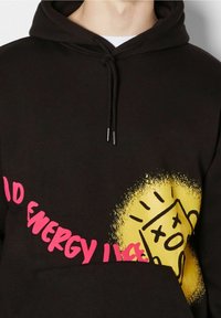 Sudadera negra con texto rosa "SIN ENERGÍA" y un gráfico estilizado de una taza de café muerta amarilla en el pecho, que cuenta con un bolsillo frontal tipo canguro.
