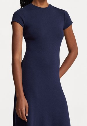 Femme portant une robe bleu marine ajustée à manches courtes, debout avec un bras détendu et l'autre plié derrière son dos.