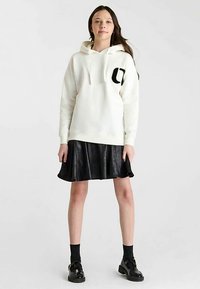 Sweat à capuche blanc avec un logo noir sur la poitrine, associé à une jupe évasée en similicuir noir et des chaussures noires.
