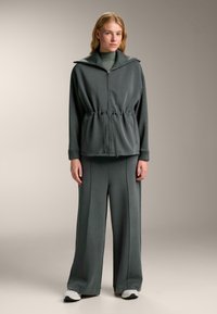 Dymovo šedý voľnočasový kostým s bundou na zips a vysokým golierom, doplnený širokými nohavicami.