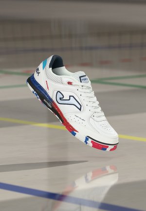 Weißer Sportschuh mit blauen, roten und schwarzen Akzenten, der über einem Hallenplatz mit grünen und gelben Linien schwebt.