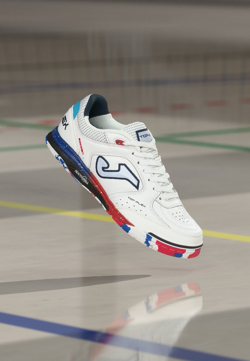 Άσπρο αθλητικό παπούτσι με μπλε, κόκκινα και μαύρα στοιχεία που αιωρείται πάνω από ένα εσωτερικό γήπεδο με πράσινες και κίτρινες γραμμές.