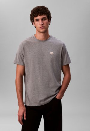 Gray short-sleeve t-shirt in soft fabric with a round neckline and a CK logo patch in beige on the left chest. Worn with dark pants.