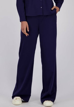 Personne portant un pantalon bleu marine ample et une chemise boutonnée assortie avec des sneakers blanches, se tenant avec une main dans la poche.