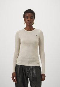 Langermet kremfarget strikket topp med rund hals, tettsittende design og liten svart logo på venstre bryst, kombinert med svarte skinnbukser.