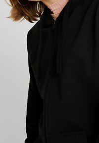 Sweat à capuche noir zippé avec une texture douce, doté d'une capuche et de cordons, ainsi que d'une poche frontale. Détails subtils le long de la fermeture éclair.