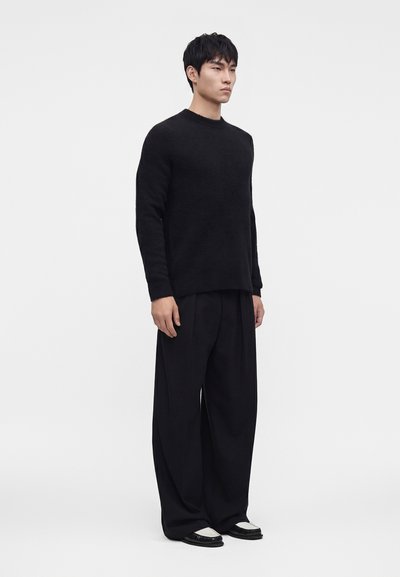 Mann steht, trägt einen schwarzen Langarmpullover, lockere schwarze Hose und schwarz-weiße Schuhe vor einem einfarbigen hellen Hintergrund.