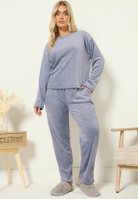 En kvinna står iklädd en ljuslila sammetsblus med lång ärm och byxor, med grå fluffiga tofflor, bredvid ett träbord med torkade växter och en stoppad stol.