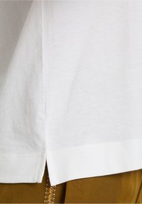 T-shirt en coton blanc avec une légère fente sur le côté à l'ourlet. Texture lisse; design décontracté. Superposé sur un tissu marron avec des accents en perles.