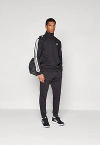 Svart atletisk träningsoverall av polyester med vita ränder på ärmarna och svarta byxor. Inkluderar en svart ryggsäck och svarta sneakers.