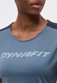 Κοντινό πλάνο ενός ατόμου που φοράει μπλε αθλητική μπλούζα με μαύρα πάνελ στους ώμους και την επιγραφή «DYNAFIT» σε λευκό χρώμα στο στήθος.