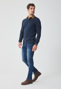 Marine langermet polo med brun krage, kombinert med mørkeblå jeans og brune snøresko. Har en liten logo på brystet.