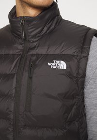 Mężczyzna noszący czarną ocieplaną kamizelkę The North Face z kieszenią na zamek błyskawiczny na piersi, pod którą ma szary longsleeve, przycięty na szyi i ramionach.