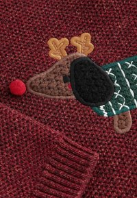 Gros plan d'un tissu tricoté rouge avec un renne brodé portant un pull vert et un nez pompon rouge.