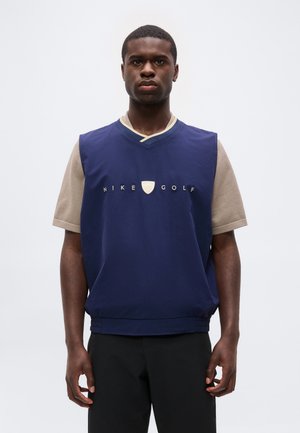 Jeune homme portant un gilet sans manches Nike Golf bleu sur une chemise beige à manches courtes, debout devant un fond blanc uni.