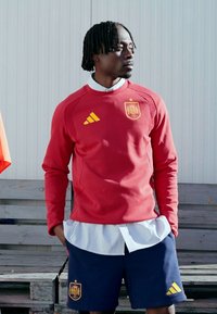 Felpa rossa con logo Adidas giallo e stemma della Spagna, abbinata a pantaloncini blu e maglietta a righe sotto, in piedi contro uno sfondo chiaro.