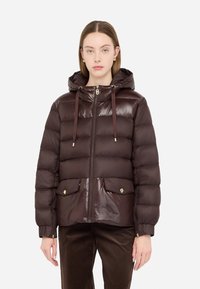 Giacca zip-up marrone, morbida e con cappuccio, finitura lucida, due tasche frontali con chiusura dorata e cordoncino di regolazione. Pantaloni di velluto a coste sotto.