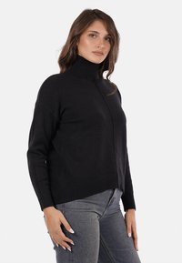 Donna che indossa un maglione nero a collo alto e jeans blu, in piedi con una mano rilassata al lato contro uno sfondo bianco.