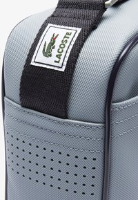 Borsa grigia con texture e una tracolla nera, dotata di etichetta con il logo Lacoste. Presenta perforazioni sui lati e cuciture delineate.