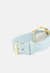 Correa de reloj de silicona azul clara con hebilla de metal dorado, que presenta una textura suave y un diseño minimalista, mostrando la parte trasera de la esfera del reloj.