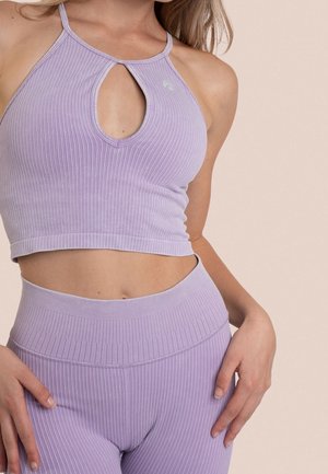 Frau trägt ein geripptes, hellviolettes bauchfreies Oberteil mit Schlüsselloch-Ausschnitt und dazu passende hoch taillierte, gerippte Leggings vor einem beigen Hintergrund.