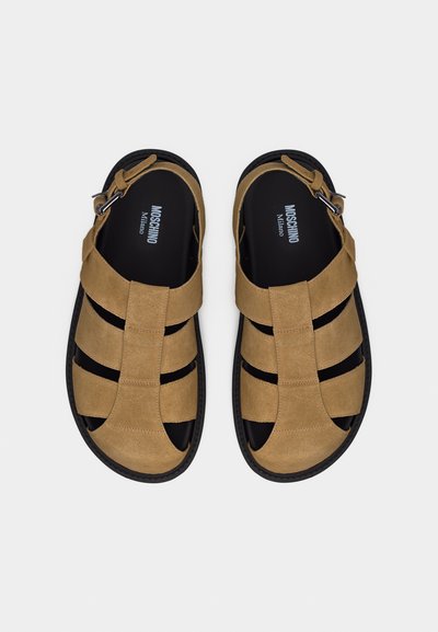 Sandales gladiateur en cuir fauve avec semelles noires et brides à boucles, portant la marque Moschino Milano sur la semelle intérieure noire, vues de dessus.