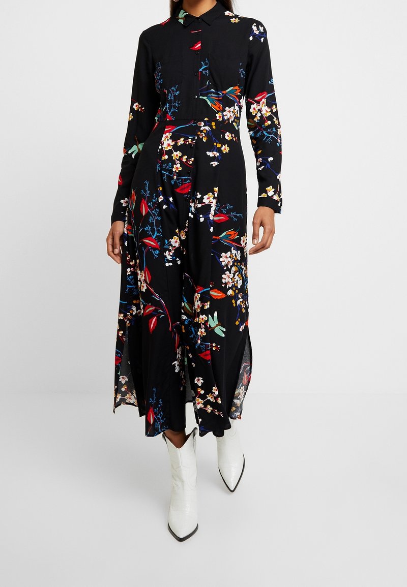 Femme portant une robe maxi noire à manches longues et motifs floraux, associée à des bottes blanches à bouts pointus, debout devant un fond uni.