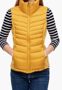 Musztardowa kamizelka puchowa z wysokim kołnierzem, poziomym pikowaniem, bocznymi kieszeniami i suwakiem z przodu. Nosi na sobie czarno-białą bluzkę w paski.