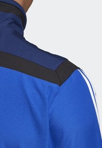 Blå idrottsjacka med hög krage, slät textur, svarta axeldetaljer och vita sidstripe för extra detalj.