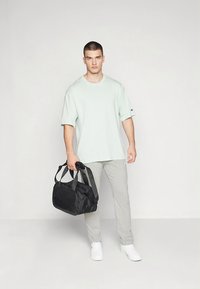 Lys grønn oversized t-skjorte, grå joggebukse, hvite joggesko og en svart duffelbag med håndtak. Enkelt, minimalistisk treningsantrekk.