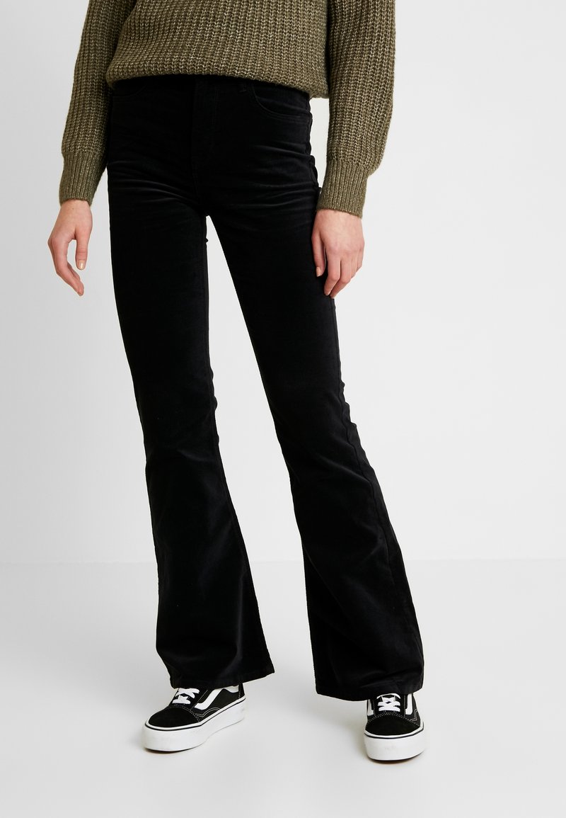 Personne portant un pantalon noir évasé, des baskets noires et blanches, et un pull en tricot vert olive, debout devant un fond uni.