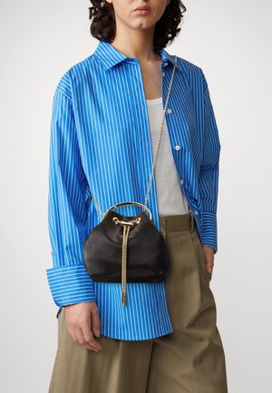 Personne portant une chemise à boutons rayée bleu et blanc par-dessus un haut blanc, un pantalon olive, et un sac à cordon noir avec une bandoulière chaîne dorée.