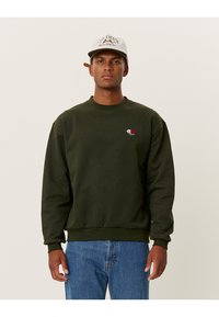 Felpa verde in cotone, con un piccolo ricamo di una rosa sul petto e polsini e orlo a costine. Indossata con jeans blu.