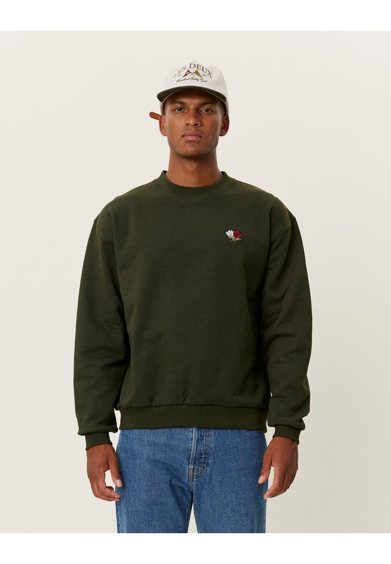 Felpa verde in cotone, con un piccolo ricamo di una rosa sul petto e polsini e orlo a costine. Indossata con jeans blu.