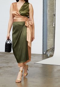 Robe portefeuille en satin présentant des teintes vert olive et cuivre, un imprimé animal, et une taille nouée. Associée à des talons dorés à brides et un sac noir.