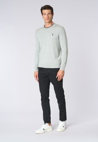 Maglione lavorato a maglia grigio chiaro con collo a costine, abbinato a jeans neri slim-fit e sneaker bianche. Il maglione presenta un piccolo logo sul petto.