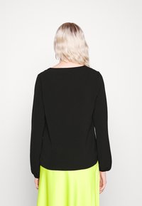 Personne blonde aux cheveux ondulés et lâches, portant un haut à manches longues noir et une jupe verte citron vif, debout face à un fond blanc.