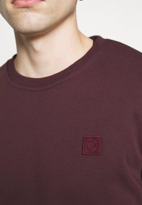 Bordowa bluza z okrągłym dekoltem wykonana z miękkiej tkaniny, z małą, haftowaną naszywką z wzorem herbu na lewej piersi.