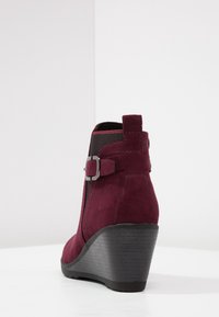 Bordeauxfarbene Wildleder-Stiefelette mit Keilabsatz, ausgestattet mit einem seitlichen Schnallenakzent, elastischen Einsätzen und einer strukturierten schwarzen Gummisohle.