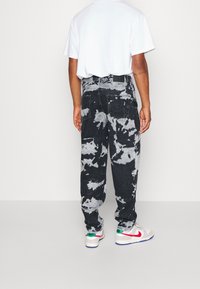 Vaqueros de mezclilla en tonos negro y gris de estilo tie-dye con un ajuste relajado, que cuentan con bolsillos traseros y una cintura alta. Llevados con zapatillas blancas con acentos de color.