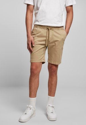 Pantalones cortos beige hasta la rodilla hechos de algodón con una cintura elástica y cordón. Se llevan con zapatillas blancas y calcetines. Diseño simple.