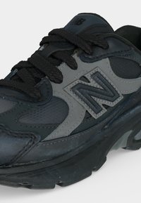 Μαύρο και γκρι αθλητικό παπούτσι New Balance με χοντρή σόλα, επενδεδυμένη γλώσσα και μαύρα κορδόνια δεμένα σε φιόγκο.