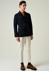 Marineblauw jas van een wolmix met twee borstzakken, een ceintuur in de taille, knoopsluiting en een hoge kraag, gecombineerd met witte taps toelopende broeken en bruine schoenen.