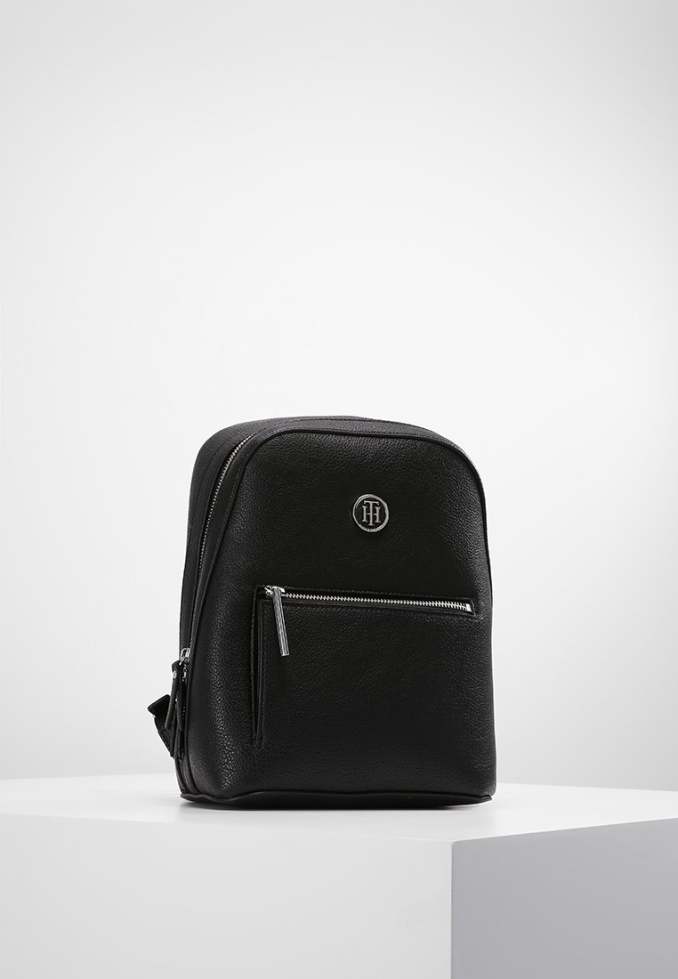 Czarny plecak z sztucznej skóry o zaokrąglonym kształcie, z przednią kieszenią na suwak i małym logo na przednim panelu. Gładka faktura, minimalistyczny styl.